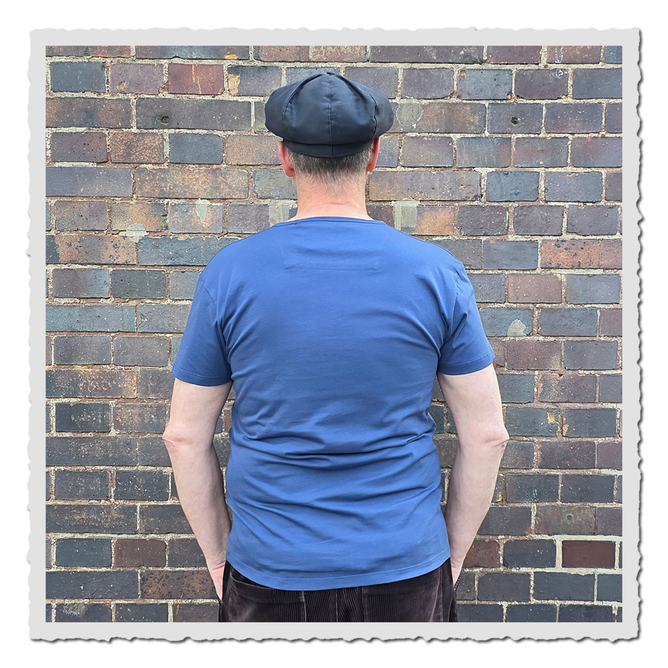 Rückansicht einer Person mit blauem T-Shirt vor einer braun-grauen Backsteinwand.